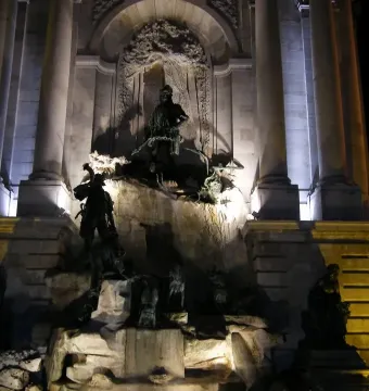 The Fountain of King Matthias illuminated at night, with dark statues on tiered rocky levels and a central figure above.