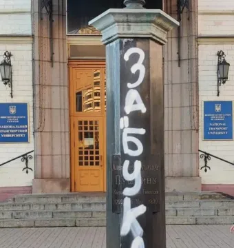 Graffiti-covered pedestal of the Pushkin Bust with a decorative cap at the top, standing before a building entrance.