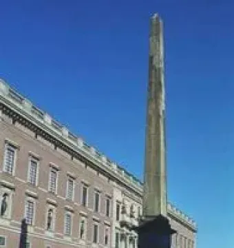 The Obelisk at Slottsbacken stands in a paved square beside a long building under a clear blue sky.