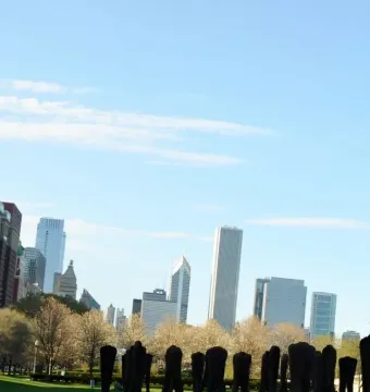 Agora sculpture in Grant Park, with dark upright figures in the foreground and the Chicago skyline beyond the trees.