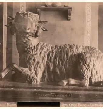 Resting bronze ram with curled horns on a long pedestal, shown against a striped and pillared wall backdrop.
