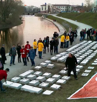 People gathered around the Keyboard Monument, with one person painting a key on the large concrete keyboard beside a river.