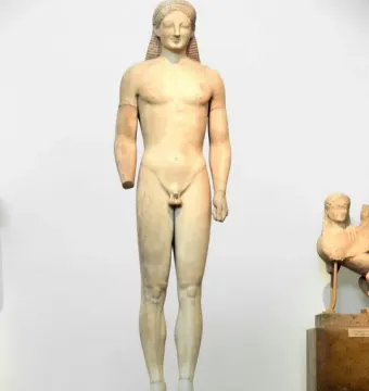 A tall marble Kouros statue of a nude male figure standing frontally with long braided hair and a missing left forearm.