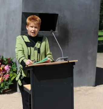 A woman speaks at a podium in front of the Memorial to Homosexuals Persecuted Under Nazism, with floral wreaths nearby.