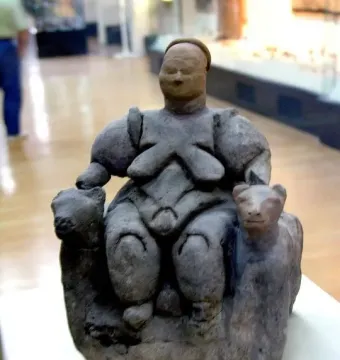 Seated Woman of Çatalhöyük figurine on a museum display surface, with a throne-like seat and animal-shaped supports.
