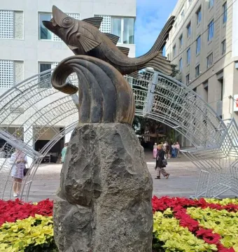 The Chicharro sculpture, a dark fish mounted on a tall stone pillar, stands in a plaza surrounded by red and yellow poinsettias.