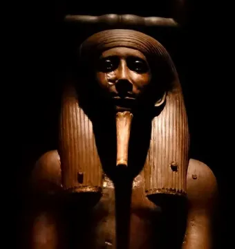 Upper body of the Ka statue of King Hor with a striped headdress and false beard against a dark background.