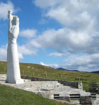 Our Lady of the Isles statue on a tiered stone platform in a grassy landscape with hills behind it