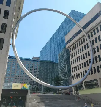 The Ring sculpture curves above a public staircase between tall city buildings.
