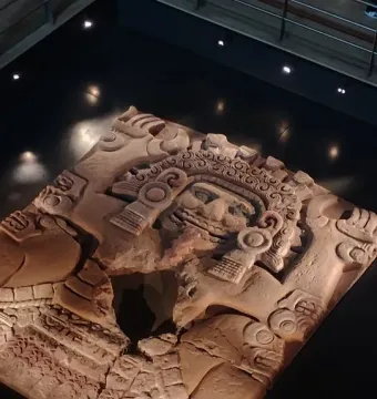 Tlaltecuhtli Monolith displayed flat in a museum, showing a carved face, headdress, and weathered stone surface.