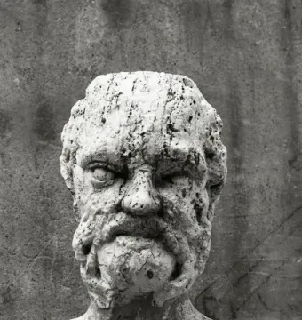 Weathered stone bust detail from the Babuino Fountain, showing a rough bearded face on a dark base.