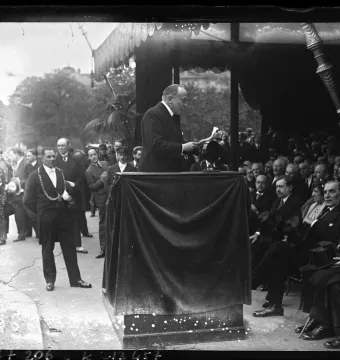 A small ornate section of the Monument to Mickiewicz is visible above a dark-draped podium as a speaker addresses a crowd.