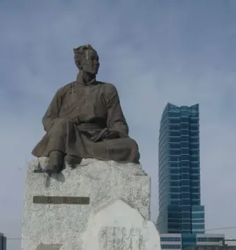 Natsagdorj Monument bronze statue on a stone pedestal with a floral wreath, with a modern glass skyscraper behind it.