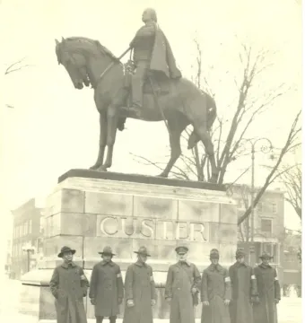 Seven uniformed men stand at attention before the Custer Monument in Monroe, Michigan, with a mounted figure atop the stone pedestal.