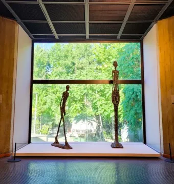 Two elongated bronze Giacometti sculptures stand on a white platform before a large window with trees outside.