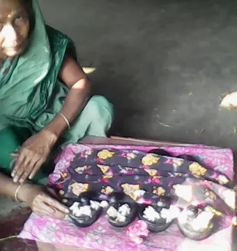 Dark padukas with toe knobs arranged on patterned cloth, with small white flowers and a woman in a green sari nearby.