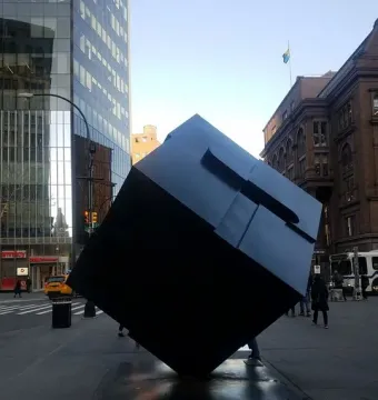 The Alamo sculpture shown as a large dark cube tilted on one corner in an urban plaza.