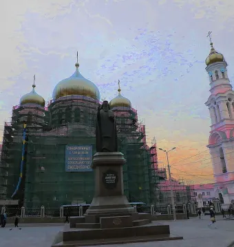 Monument to Dimitry of Rostov on a tiered pedestal in a square, with a scaffolded church, bell tower, and sunset sky behind it.