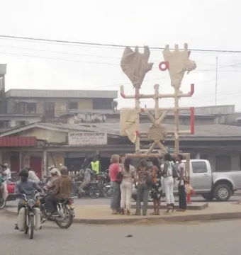 Njé Mo Yé, a tall abstract sculpture on a street median with light-colored beams, red accents, and stylized upper forms, surrounded by pedestrians and traffic.