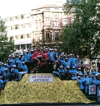 A group of people in blue shirts and white hats gathered around a yellow, banana-patterned sculpture covered with a tarp.