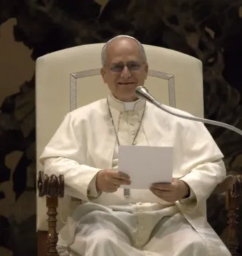 A smiling pope in white clerical vestments, glasses, and a skullcap speaks into a microphone while holding a blank paper.