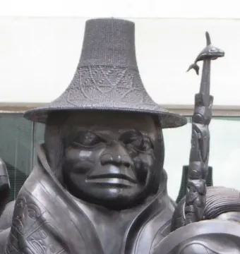 Close-up of Spirit of Haida Gwaii showing a carved figure in a woven-textured hat and patterned cloak beside a whale-topped staff.