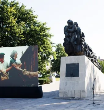 Train of Pain monument on a light stone base in a paved square, with a large historical display screen beside it.