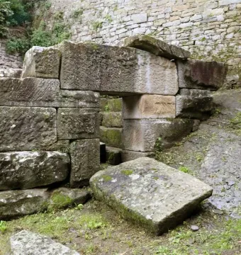 An Etruscan tomb with a small rectangular doorway in a moss-covered ashlar structure, with a fallen stone on the grass in front.