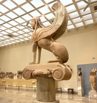 The Sphinx of Naxos shown in left profile on a fluted column inside a museum hall.