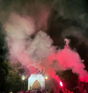The Statue of Honor stands on a pedestal at night as red and white smoke rises around a crowd below.