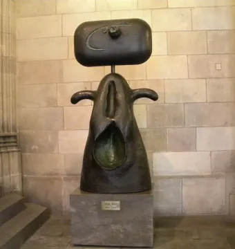 Femme, a dark abstract sculpture with a rectangular head-like top and curved forms, displayed on a stone pedestal indoors.