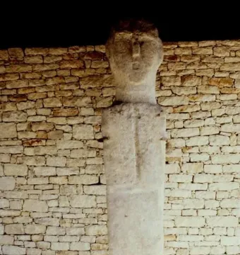 U Nativu, a pale stone figure with an elongated head and simple facial features stands against a dry-stone wall.
