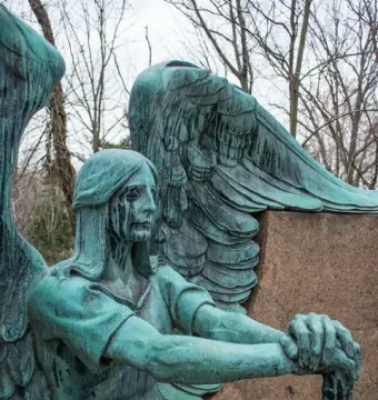 Left detail of the Haserot Angel, showing a weathered greenish winged figure holding a staff with dark streaks on its face.