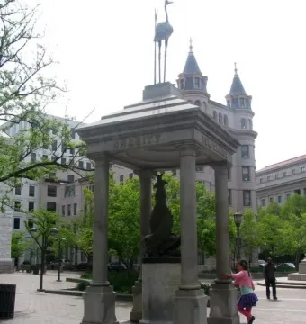 Temperance Fountain with a bird figurine on top in a paved urban plaza, with a girl standing nearby and a man walking in the background.