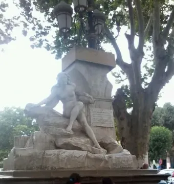 Ain El Fouara Fountain with a reclining nude female statue on a rocky base and a lamppost above it, framed by a leafy tree.