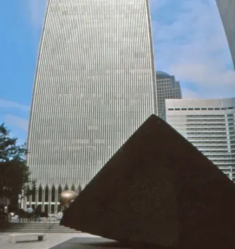Cloud Fortress, a dark triangular sculpture on a paved plaza with a skyscraper rising behind it.