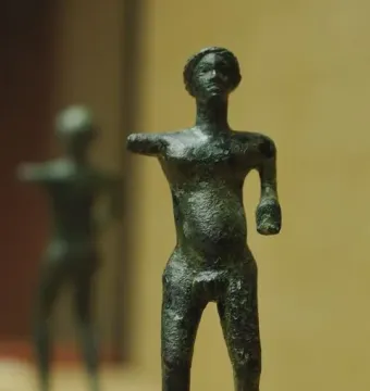 Bronze standing nude male statuette from the Neuvy-en-Sullias Hoard on a marbled base, with a similar figure blurred in the background.