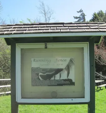 Informational kiosk for Running Fence stands on a grassy lawn near a split-rail fence.
