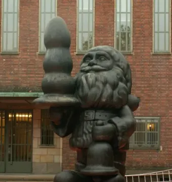Dark Santa Claus statue holding a bell and a large black bulbous object in front of a brick building.