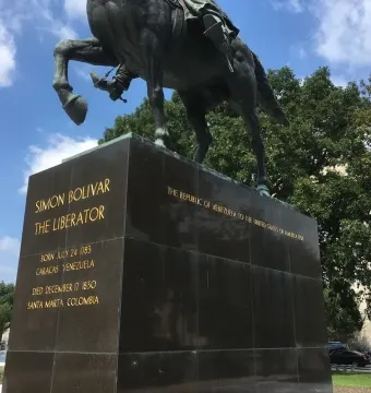 Lower section of the Simón Bolívar equestrian statue on a dark stone pedestal with gold inscriptions, framed by trees and a cloudy blue sky.