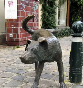Zinneke Pis bronze dog sculpture on a cobblestone street beside a dark green bollard.