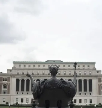 Alma Mater bronze statue seen from behind with Columbia University buildings and columns in the background.
