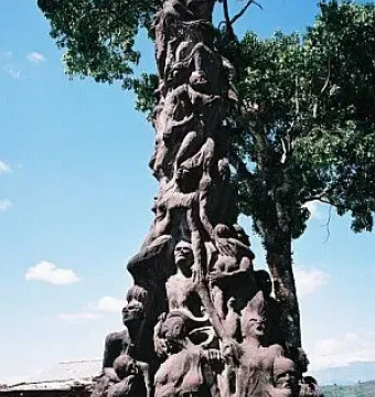 Pillar of Shame sculpture with writhing human figures on a tall dark pillar and plaque at the base.