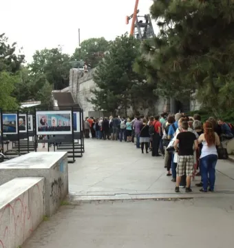 A long queue of people stands below the Prague Metronome near display panels and trees.