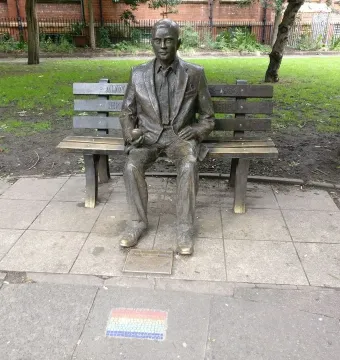 Alan Turing Memorial bronze statue seated on a bench holding an apple, with a mosaic foreground and park behind it.