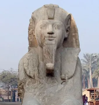 The Sphinx of Memphis, a weathered stone sphinx with a pharaonic headdress and false beard, stands on a rough base amid palm trees.