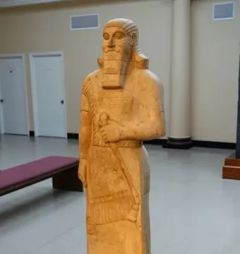 Statue of Ashurnasirpal II standing on a pedestal in a museum gallery, with a bearded figure holding an object across his chest and wearing a long patterned tunic and fringed skirt.