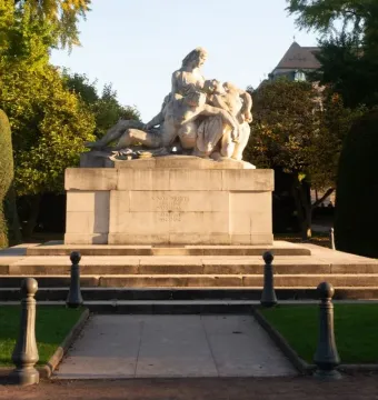 The Strasbourg War Memorial, a large stone sculpture of figures in a mournful embrace on a tiered pedestal, surrounded by grass, bushes, and autumn trees.
