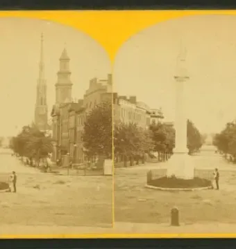 Abraham Lincoln monument on a tall column in the middle of a street, with a small figure near its fenced base and buildings in the background.