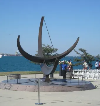 A large dark sculptural sundial stands on a circular stone base beside the water with boats in the distance.